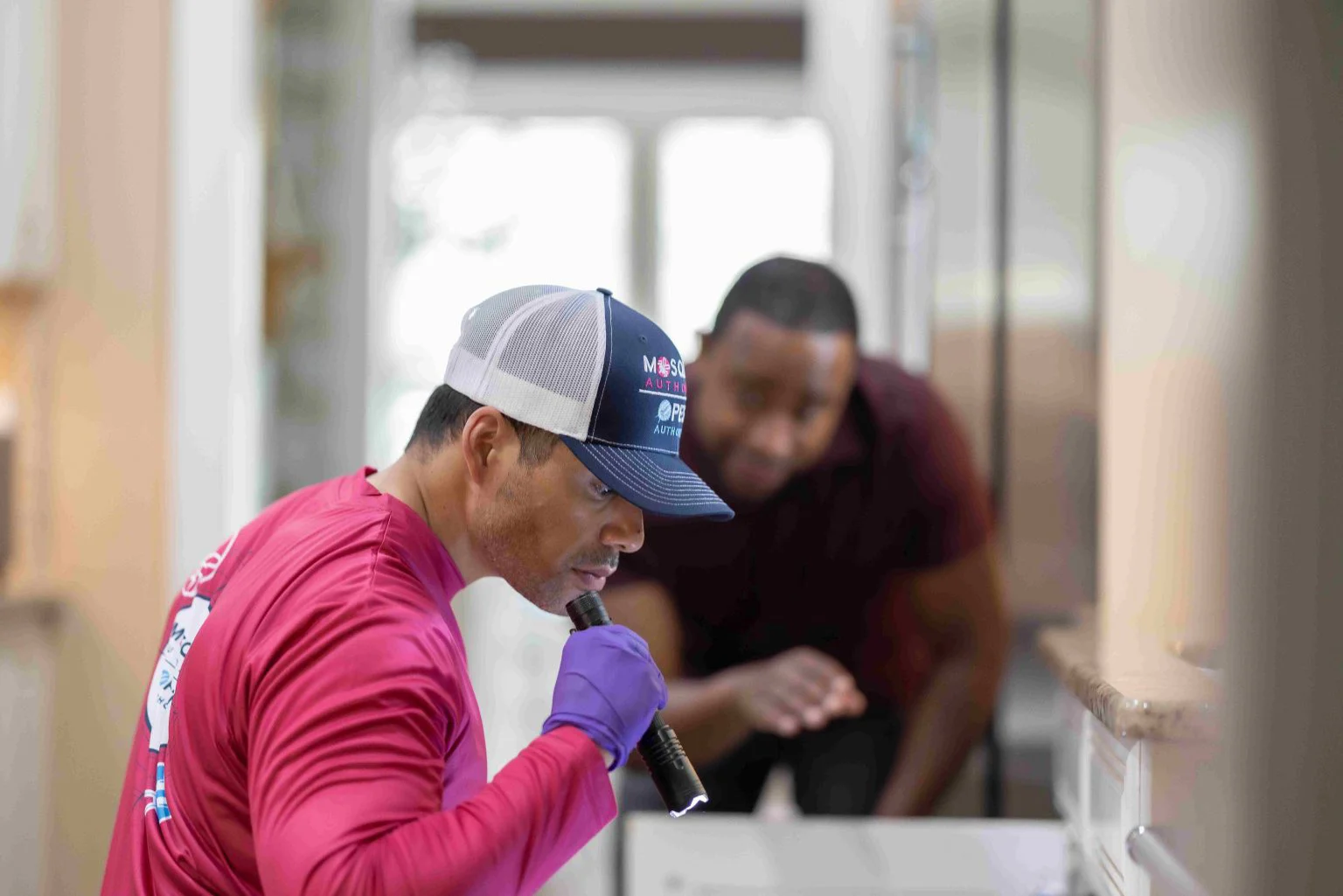 A pest control technician in a branded cap and gloves meticulously inspects an area with a flashlight